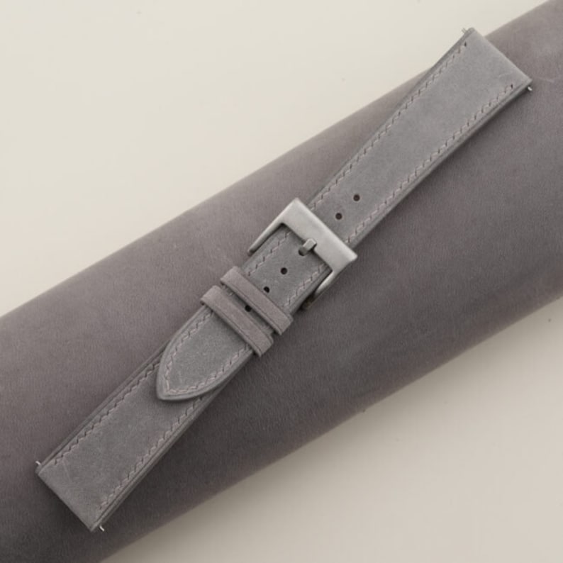 May include: A gray suede watch strap with a silver buckle. The strap is rolled up on a gray surface.