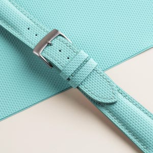 Puede incluir: Primer plano de una correa de reloj azul claro con una superficie texturizada y una hebilla plateada. La correa est&aacute; cosida con hilo azul claro y se encuentra sobre una superficie texturizada a juego.