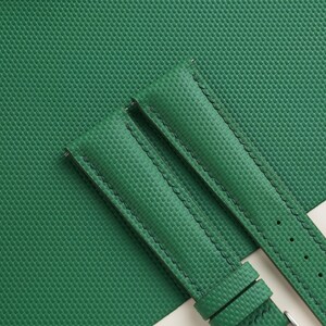 May include: Close-up of a green watch strap with a textured surface and black stitching. The strap is made of a durable material and features a buckle. The background is a matching green surface.