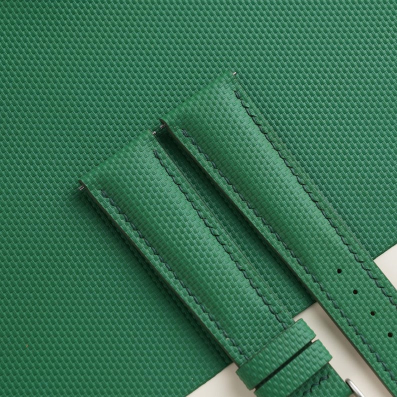 May include: Close-up of a green watch strap with a textured surface and black stitching. The strap is made of a durable material and features a buckle. The background is a matching green surface.