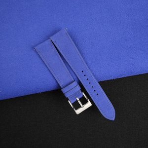 May include: Two blue suede watch straps with a silver buckle. The straps are laid out on a blue and black surface.