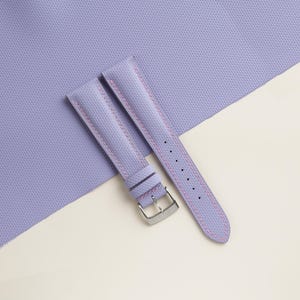 Puede incluir: Una correa de reloj lavanda con costuras rosas y una hebilla plateada. La correa est&aacute; hecha de un material texturizado y se muestra sobre una superficie lavanda y crema. La correa tiene varios agujeros para ajustar.