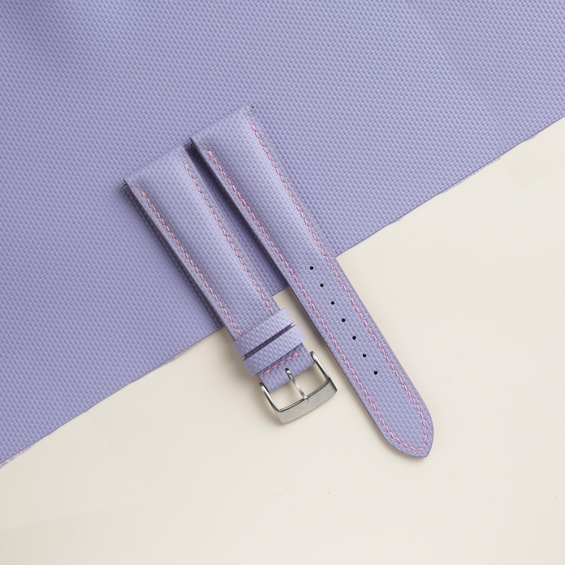 Puede incluir: Una correa de reloj lavanda con costuras rosas y una hebilla plateada. La correa est&aacute; hecha de un material texturizado y se muestra sobre una superficie lavanda y crema. La correa tiene varios agujeros para ajustar.