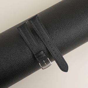 May include: Two black leather watch straps with a silver buckle. The straps are rolled up and placed on a black textured surface.