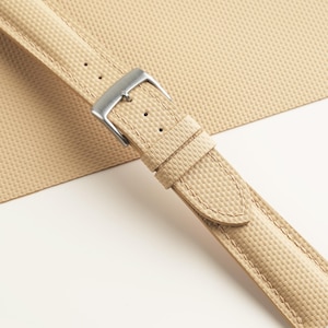 Puede incluir: Correa de reloj beige con hebilla de acero inoxidable. La correa tiene un patr&oacute;n de tejido texturizado y est&aacute; cosida a lo largo de los bordes. El fondo es de color beige a juego.