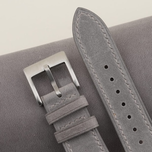 May include: A gray leather watch strap with a silver buckle. The strap has stitching around the edges and is made of a soft, suede-like material.