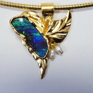 Könnte beinhalten: Eine goldene Halskette mit einem Anhänger aus einem blauen und grünen Opal, der in ein Blattdesign aus Gold gefasst ist. Der Anhänger ist mit drei kleinen weißen Perlen verziert.