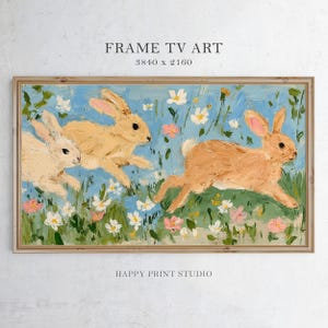 以下が含まれることがあります： 3匹のウサギが花畑を駆け抜ける様子を描いた額入りの絵画。淡い青色の背景に白とピンクの花が咲いています。「FRAME TV ART」と「HAPPY PRINT STUDIO」の文字が見えます。