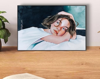 May include: A watercolor painting of a young person with brown hair, wearing a white shirt, lying on a bed with a blue and green background. The painting is framed in a black frame.
