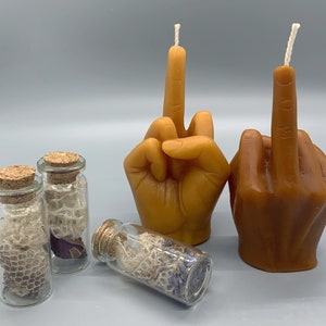 K&ouml;nnte beinhalten: Zwei braune handf&ouml;rmige Kerzen mit Dochten, jede mit einem erhobenen Mittelfinger in einer unh&ouml;flichen Geste. Drei kleine Glasgef&auml;&szlig;e mit Korkdeckeln enthalten getrocknete Blumen und Spitze.