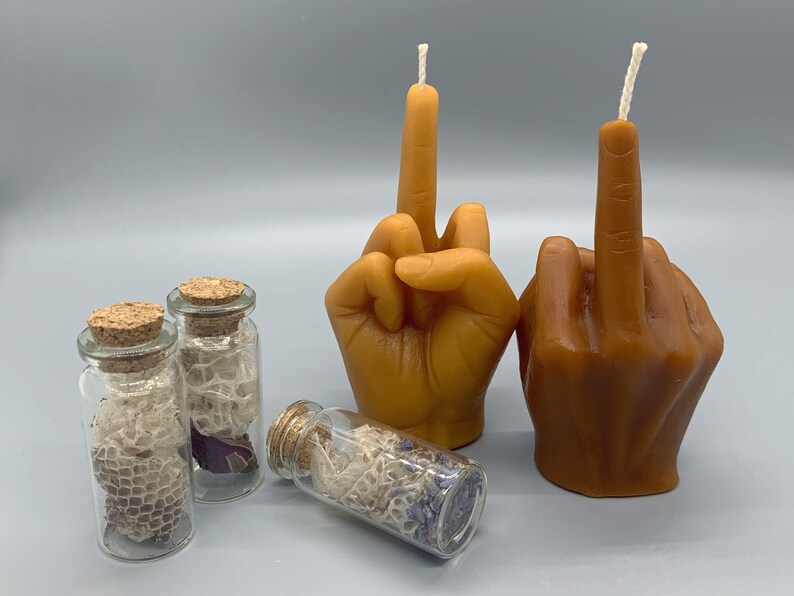 K&ouml;nnte beinhalten: Zwei braune handf&ouml;rmige Kerzen mit Dochten, jede mit einem erhobenen Mittelfinger in einer unh&ouml;flichen Geste. Drei kleine Glasgef&auml;&szlig;e mit Korkdeckeln enthalten getrocknete Blumen und Spitze.