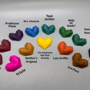 K&ouml;nnte beinhalten: Eine Sammlung von 12 herzf&ouml;rmigen Wachs-Schmelzen in verschiedenen Farben, darunter Rot, Orange, Gelb, Gr&uuml;n, Blau, Lila und Schwarz. Jede Schmelze hat einen anderen Namen darunter, wie "Professor Plum", "Teal Jerker", "The Simpsons Did That Already" und "Lois Griffin".