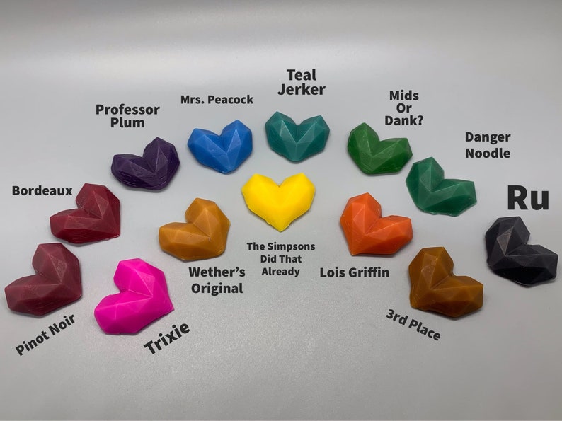 K&ouml;nnte beinhalten: Eine Sammlung von 12 herzf&ouml;rmigen Wachs-Schmelzen in verschiedenen Farben, darunter Rot, Orange, Gelb, Gr&uuml;n, Blau, Lila und Schwarz. Jede Schmelze hat einen anderen Namen darunter, wie "Professor Plum", "Teal Jerker", "The Simpsons Did That Already" und "Lois Griffin".