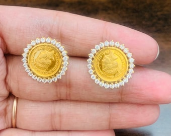 May include: A pair of gold-toned stud earrings. Each earring features a circular coin design with a portrait and text, surrounded by a halo of small, clear gemstones. The earrings are set against a neutral background.