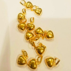 Puede incluir: Un grupo de 11 colgantes de corazón de oro con un bucle en la parte superior para sujetarlos a una cadena. Los colgantes están dispersos sobre una superficie blanca.