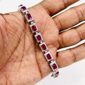 Op de afbeelding: Een zilveren armband met rode edelstenen in een vierkante vorm. De armband is gemaakt van metaal en heeft een gepolijste afwerking.