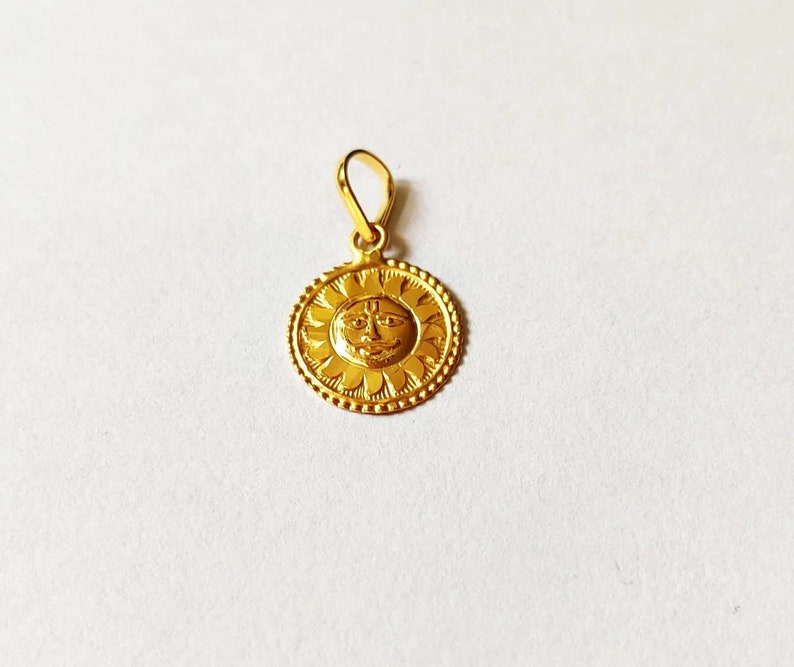 May include: A gold pendant featuring a sun with a face in the center. The sun is surrounded by a circle of small flowers.