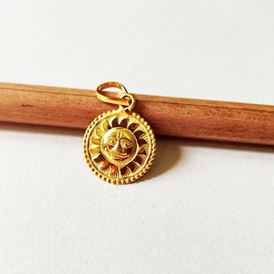 May include: A gold pendant featuring a sun with a face, surrounded by a decorative border of rays. The pendant is on a wooden surface.