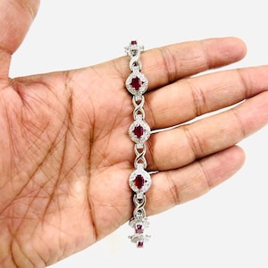 Op de afbeelding: Een zilveren armband met een kettinglinkontwerp en rode ovale edelstenen die zijn gezet in een halo van kleinere witte edelstenen.