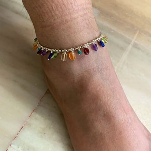 May include: A silver anklet with a chain design, adorned with colorful gemstone beads in various shapes. The gemstones include orange, yellow, green, blue, purple, and red. The anklet is worn on an ankle.