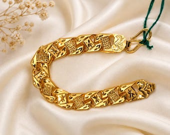 May include: A gold chain bracelet featuring alternating rectangular links with textured, cross-hatched surfaces and polished, engraved floral patterns. The bracelet includes a gold hook clasp with a green string attached to one end of the metal chain.