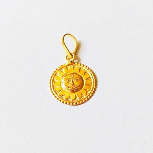 May include: A gold pendant featuring a sun with a face and a "U" symbol in the center. The pendant is on a white background.