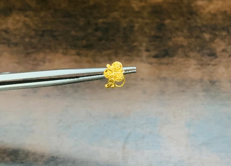 May include: A gold nose ring with a flower design. The flower has five petals, each with a textured surface.