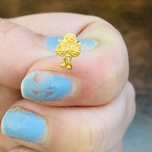 May include: A gold nose ring with a floral design. The ring has three round flowers with a small dangling charm.