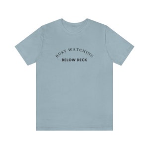 Op de afbeelding: Een lichtblauw t-shirt met de tekst "Busy Watching Below Deck" erop gedrukt.