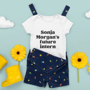 May include: White baby onesie with black text that reads "Sonja Morgan's future intern". The onesie is paired with blue denim overalls with a colorful pattern.