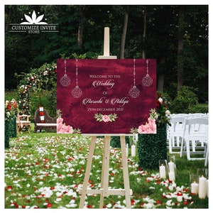 May include: A maroon wedding welcome sign with white text and silver glitter accents. The sign reads "Welcome to the Wedding of Aarushi & Adilya 25th December 2021".