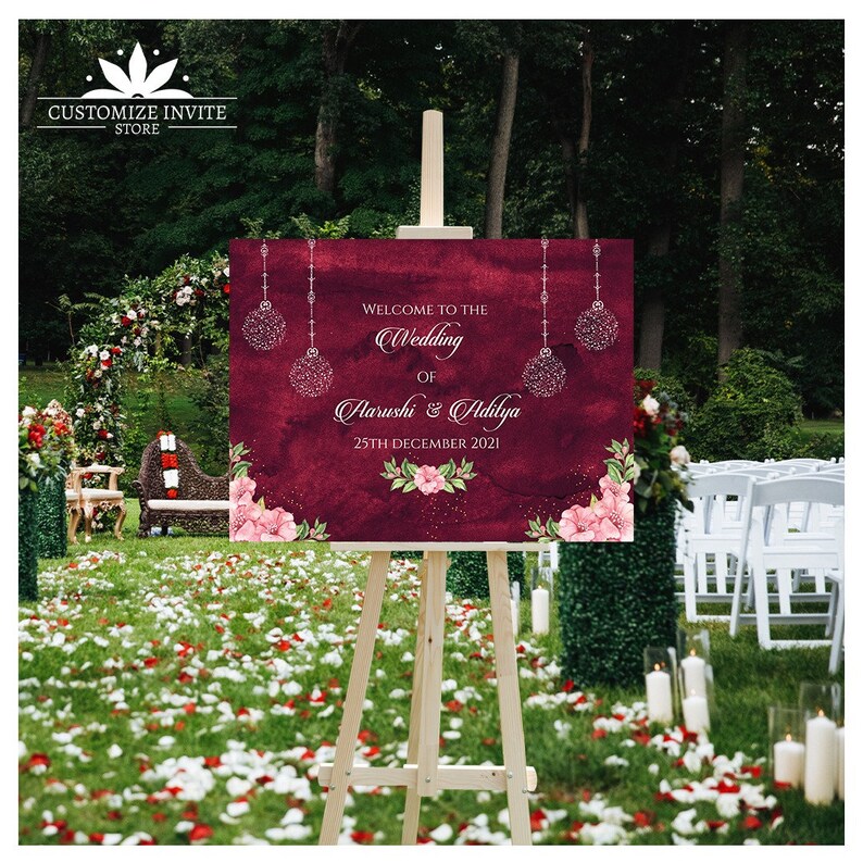 May include: A maroon wedding welcome sign with white text and silver glitter accents. The sign reads "Welcome to the Wedding of Aarushi & Adilya 25th December 2021".