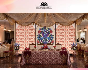 May include: A wedding reception setup featuring a central table with a dark patterned tablecloth, positioned in front of three decorative panels with floral motifs. The backdrop includes draped beige fabric, hanging lanterns, and floral arrangements.