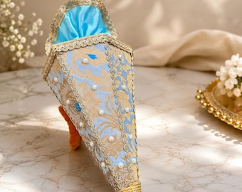 May include: A cone-shaped decorative holder featuring a light blue and gold damask fabric pattern, accented with white faux pearls, gold lace trim, and a bright blue satin interior lining, finished with a gold wrapped tip at the bottom.