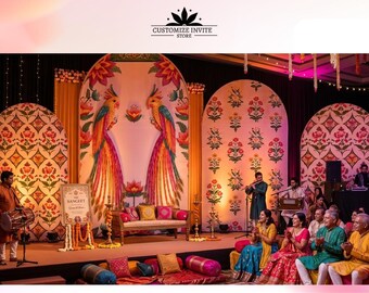 May include: A stage backdrop featuring arched panels with floral patterns and two large colorful parrots. A wooden bench sits in the center, flanked by musicians and guests in traditional attire. A sign reads THE SANGEET and CUSTOMIZE INVITE STORE.