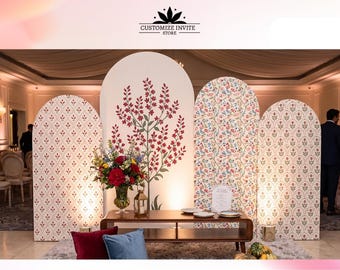 May include: Four arched event backdrops featuring cream backgrounds with repeating floral patterns and a central tree illustration. A wooden coffee table sits in front with a floral arrangement, a gold lantern, and two solid-colored throw pillows.