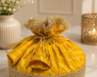 May include: A mustard yellow drawstring pouch featuring a white wavy line pattern, a gold metallic braided trim along the base, and a ruffled top edge finished with gold metallic thread. The bag is cinched with matching gold cord drawstrings.