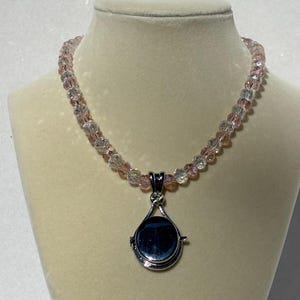 May include: A beaded necklace with a silver pendant. The necklace features small, faceted beads in shades of pink and clear. The pendant is a teardrop shape with a silver frame and a dark, reflective center. A small, clear gem is set at the top of the pendant.