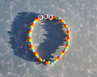 Könnte beinhalten: Ein farbenfrohes Perlenarmband mit den Buchstaben "OBX", die in weißen Perlen geschrieben sind. Das Armband besteht aus orangefarbenen, gelben und blauen Perlen.