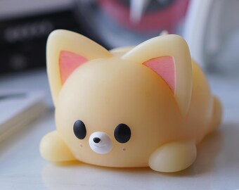 May include: A small, yellow, plush toy shaped like a fox with pink ears and black eyes. The toy is lying on a white surface.