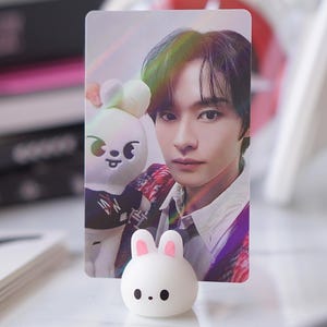 May include: A white photo card with a holographic effect features a young man with dark hair and a white shirt. He is holding a white plush toy with black eyes and a pink nose. The photo card is propped up by a white bunny-shaped stand with pink ears.