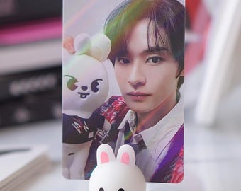 May include: A white photo card with a holographic effect features a young man with dark hair and a white shirt. He is holding a white plush toy with black eyes and a pink nose. The photo card is propped up by a white bunny-shaped stand with pink ears.