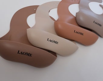 Puede incluir: Cuatro baberos de silicona en diferentes tonos de marrón con un recogedor de comida integrado. Cada babero tiene la palabra "LAOME" impresa.