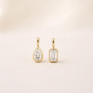 May include: Two gold pendants with diamond gemstones. One pendant features a pear-shaped diamond, and the other has an emerald-cut diamond. Both pendants have a simple, elegant design.
