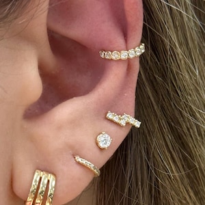 May include: Close-up of an ear adorned with multiple gold earrings. The ear features a curved ear cuff with small, clear stones, a bar earring, a stud, and a triple hoop earring. The jewelry is set against the backdrop of the person's skin and hair.
