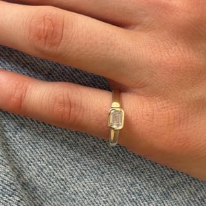 May include: A gold ring featuring a single emerald-cut clear gemstone set in a bezel setting. The band is smooth and polished, showcasing a clean and simple design that highlights the rectangular shape of the center stone on a finger.