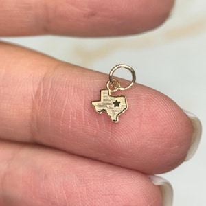 May include: A small gold-colored charm in the shape of the state of Texas, with a star detail. The charm is attached to a small ring. The charm is held between two fingers.