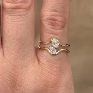 May include: A gold bypass ring featuring two distinct bands that curve around the finger. The ring is set with two clear, faceted stones: one oval-shaped stone on the upper band and one pear-shaped stone on the lower band, creating a balanced design.