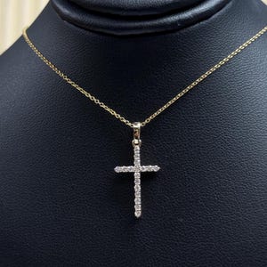 Puede incluir: Un collar dorado con un colgante de cruz. La cruz está adornada con pequeñas piedras preciosas transparentes. El collar se presenta sobre un fondo negro.