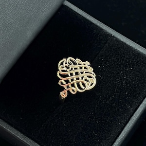 以下が含まれることがあります： 黒いジュエリーボックスに入った、複雑な織り模様のゴールドカラーのリング。 リングの模様は、独特で質感のある外観を作り出す、一連の絡み合った線です。 リングはジュエリーです。